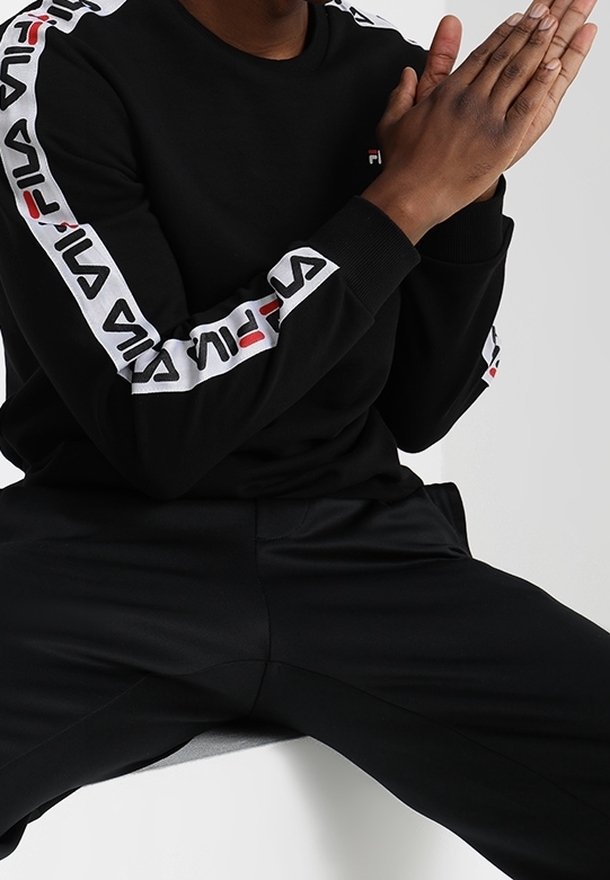 Chemise noire à manches longues avec des bandes de logo FILA blanches et rouges le long des manches, associée à un pantalon noir. Tissu lisse et coupe décontractée.