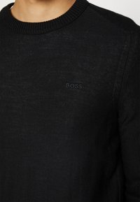 Gros plan sur un pull noir en maille avec un col côtelé et un logo "BOSS" subtil brodé sur la poitrine.