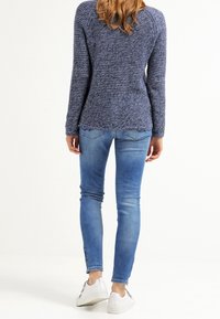 Femme debout de dos, portant un pull bleu texturé, un jean skinny bleu clair et des baskets blanches sur un fond blanc.
