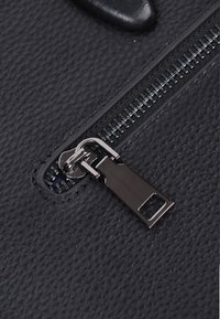 Sort tekstureret læder med en glat lynlås, der har en metallisk trækstrop og tænder. Nærbillede af lynlåsen på overfladen.