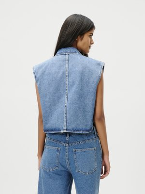 Γυναίκα με μακριά μαύρα μαλλιά, που φοράει αμάνικο γαλάζιο τζιν γιλέκο και τζιν παντελόνι με ψηλή μέση, στραμμένη πλάτη, σε απλό φόντο.