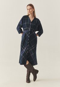 Tengerészes kockás ruha puha anyagból, V-nyakkal, hosszú ujjakkal, gombos előlappal és középhosszú szoknyával. Barna térdig érő csizma.