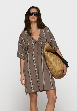 Kobieta w za dużej, pasiastym sukience i dużych czarnych okularach przeciwsłonecznych, trzymająca plecioną słomianą torbę na tle jednolitego tła.