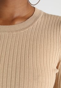 Haut en tricot côtelé beige avec un col rond, présentant des lignes texturées. La vue en gros plan met en valeur les détails de la chemise et le tissu.