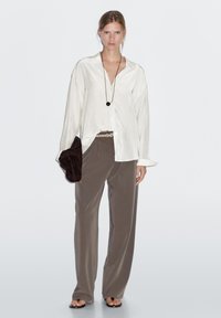 White satin shirt with a loose fit, brown wide-leg trousers, dark brown clutch, and a long black pendant necklace. Smooth texture throughout.
