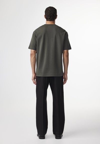 Mörkgrön kortärmad t-shirt med rak skärning, i kombination med svarta vida byxor, sedd bakifrån. Slät textur, enkel design.