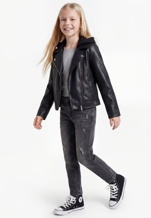 Ragazza sorridente con lunghi capelli biondi che indossa una giacca di pelle nera, jeans grigi strappati e sneakers nere alte che cammina su sfondo bianco.