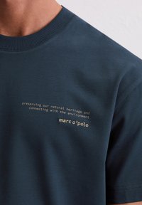 Dunkelblaues Baumwoll-T-Shirt mit geripptem Kragen, das weißen Text trägt: "Unser Naturerbe bewahren und sich mit der Umwelt verbinden."