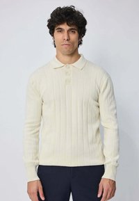 Homme aux cheveux bouclés et foncés, portant un polo à manches longues en tricot côtelé crème et un pantalon foncé, se tenant devant un fond clair uni.
