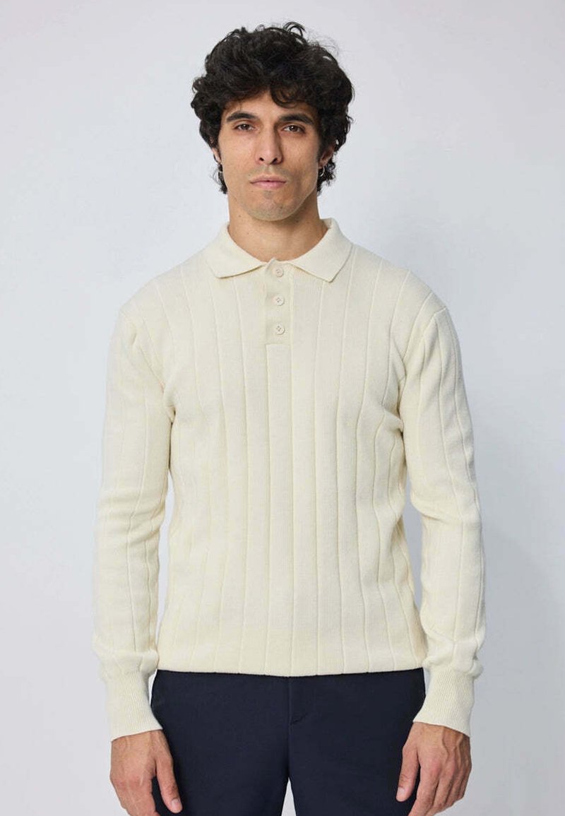 Homme aux cheveux bouclés et foncés, portant un polo à manches longues en tricot côtelé crème et un pantalon foncé, se tenant devant un fond clair uni.