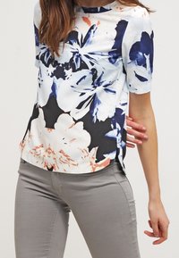 Top floreale a maniche corte bianco, blu navy e corallo. Tessuto liscio con uno sfondo nero e spacchi laterali. Abbinato a pantaloni grigi chiari.