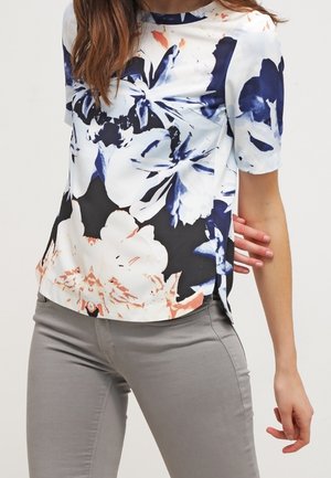 Top floreale a maniche corte bianco, blu navy e corallo. Tessuto liscio con uno sfondo nero e spacchi laterali. Abbinato a pantaloni grigi chiari.