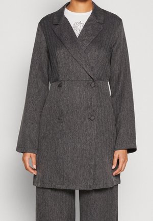 Kobieta w ciemnoszarej dwurzędowej marynarce z dopasowanymi spodniami i białym wzorzystym topem pod spodem.