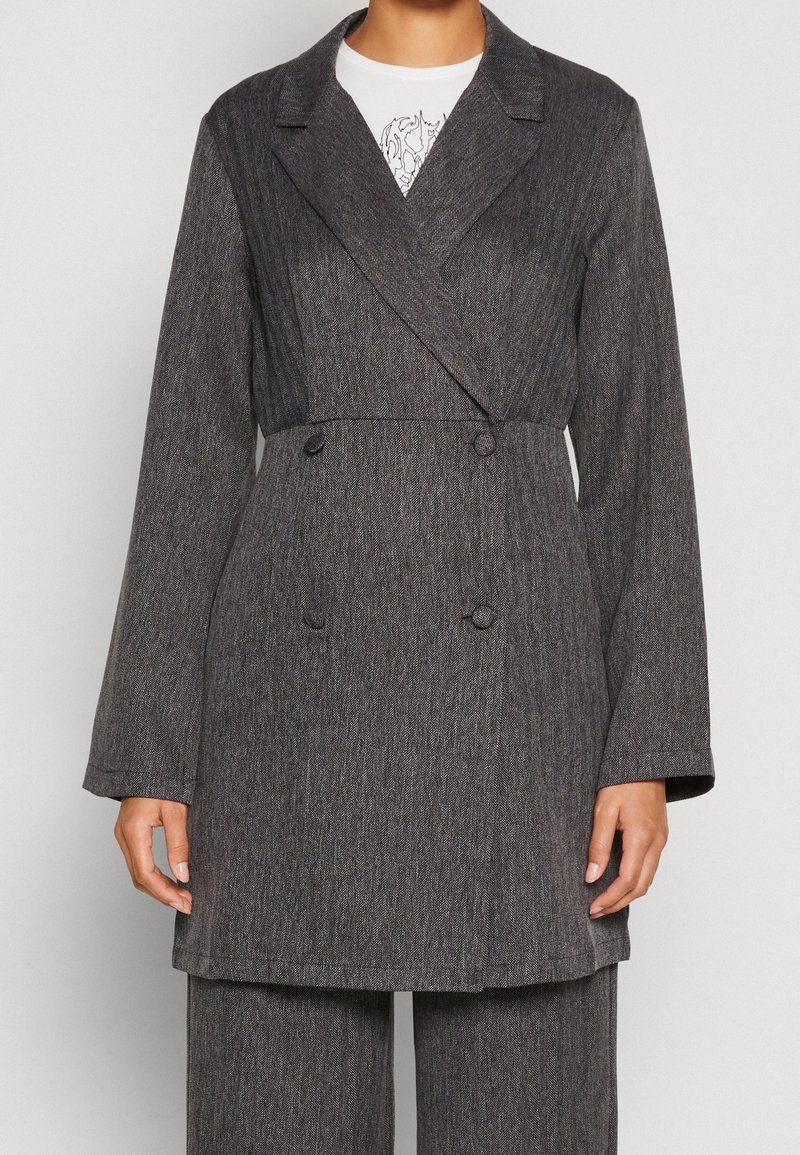 Femme portant un blazer double boutonnage gris foncé avec un pantalon assorti et un top blanc à motifs en dessous.