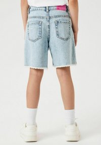 Shorts en denim bleu clair avec un ourlet effiloché, deux poches arrière et une étiquette rose à la taille. Associés à des baskets blanches et des chaussettes.