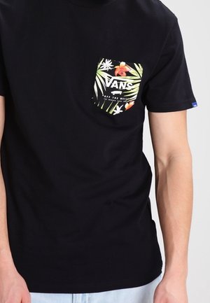 Czarna koszulka z krótkim rękawem z kolorowym, kwiatowym logo Vans na kieszeni po lewej stronie klatki piersiowej, noszona przez osobę o jasnej karnacji i w jasnoniebieskich dżinsach.