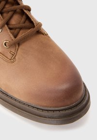 Botte en cuir marron avec bout rond, lacets marron, œillets en métal et semelle épaisse cousue sur fond gris clair.