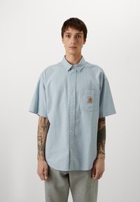 Chemise à manches courtes bleu clair avec un tissu texturé, fermeture à boutons à l'avant et une poche sur la poitrine gauche avec une étiquette de marque.