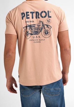 Hombre con camiseta color melocotón con gráfico de motocicleta vintage en azul oscuro y texto "PETROL, U.S.A. Outdoor Supply Florida" en la espalda, combinado con vaqueros azules.