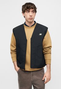 Νεαρός άνδρας με σγουρά μαλλιά, φορώντας μαύρο αμάνικο γιλέκο πάνω από μια μουσταρδί φούτερ και καφέ παντελόνι, στέκεται με τα χέρια στις τσέπες.