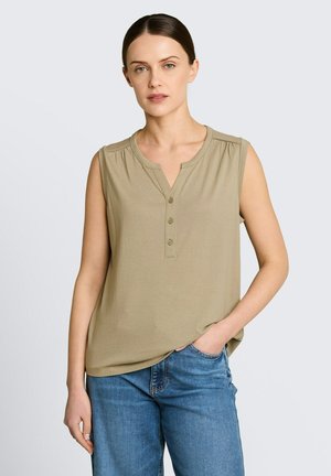 Kobieta o brązowych włosach związanych z tyłu, ubrana w oliwkowy bezrękawnik zapinany na guziki i niebieskie dżinsy, stojąca z jedną ręką w kieszeni.