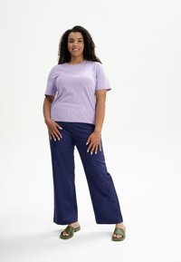 Lila kurzärmliges T-Shirt kombiniert mit marineblauen weit geschnittenen Hosen. Das Modell trägt grüne Rutsch-Sandalen. Glatter Stoff mit lockerer Passform.