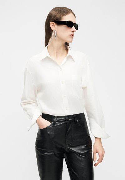 Mulher a usar óculos de sol retangulares pretos, camisa branca com botões, brincos longos de cristal pendentes e calças de couro pretas de cintura alta.