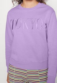 Persona che indossa una felpa lilla con "TOKYO" ricamato sulla parte anteriore e pantaloni a righe multicolori.