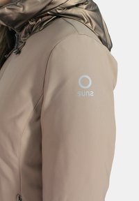 Giacca imbottita beige con cappuccio lucido marrone, caratterizzata da un logo argentato sulla manica sinistra e una texture del tessuto liscia. Dettaglio con chiusura a zip.