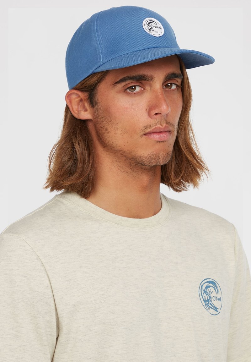 Blå kasket med en flad skygge og et cirkulært hvidt logo foran. Båret med en lysegrå t-shirt med et blåt grafisk design.