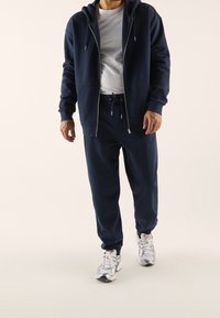 Felpa con cappuccio con cerniera blu navy con coulisse, abbinata a pantaloni jogger coordinati. Entrambi realizzati in tessuto morbido, modello indossa scarpe sportive argentate.