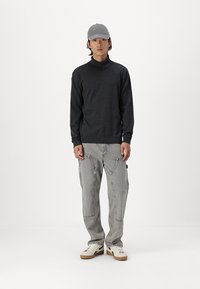 Grå langærmet turtleneck, grå lige bukser, hvide sneakers med beige accenter og en grå kasket båret af en person, der står foran en hvid baggrund.