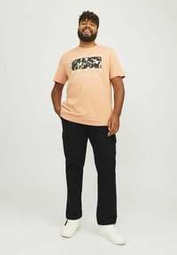 Perzikkleurig T-shirt met korte mouwen, voorzien van een zwart grafisch ontwerp en tekst, gecombineerd met zwarte cargobroeken en witte sneakers. Soepele stof, relaxte pasvorm.