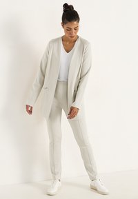 Ljusgrå blazer och matchande byxor med en slät yta, långa ärmar, sidodragkedjor och en figurnära design. Matchas med vita sneakers.