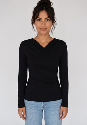 Donna con capelli scuri raccolti in uno chignon, indossa un top nero a maniche lunghe avvolgente e jeans azzurri chiari su uno sfondo chiaro uniforme.
