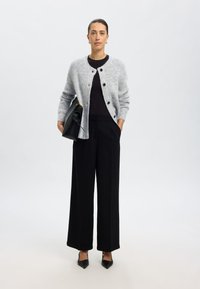 Grey, fluffy cardigan with round neck and black buttons, paired with black wide-leg trousers and pointed black heels. Holding a black bag.
