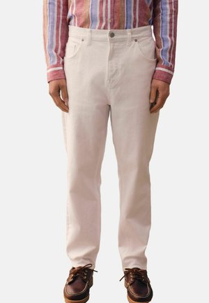 Muž nosiaci biele nohavice s rovným strihom, hnedé kožené šnurovacie topánky a košeľu s dlhým rukávom s vertikálnymi viacfarebnými pruhmi.