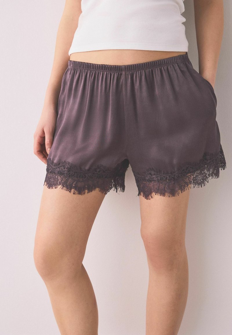 Donna che indossa pantaloncini larghi viola scuro con bordi in pizzo nero, abbinati a una canotta bianca senza maniche, in piedi contro uno sfondo neutro.