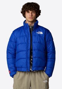 Giacca a vento blu isolata con colletto alto, apertura con zip e accenti del logo. Il tessuto appare morbido e lussuoso con un design a trapunta.