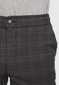 Pantaloni a quadri grigi con chiusura a bottone, vita elastica e motivi sottili in tonalità di blu scuro e marrone.