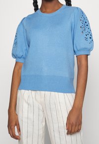 Maglione lavorato a maglia di un azzurro chiaro con maniche corte e a sbuffo, decorate con ritagli a forma di fiore. Collo a coste e orlo inferiore, indossato con pantaloni a righe.