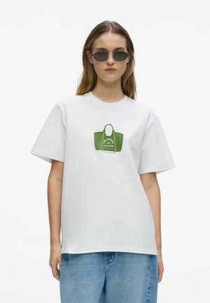 Mujer con gafas de sol, camiseta blanca con gráfico de bolso verde y vaqueros azul claro, posando sobre fondo blanco liso.
