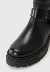 Zwarte leren laars met een gladde afwerking, voorzien van een brede riem met metalen hardware en een dikke, gestructureerde zool voor grip.