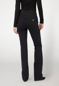 Jean noir taille haute en denim avec une légère évasement, présentant des poches arrière doubles et un logo en triangle sur la ceinture. Texture lisse.