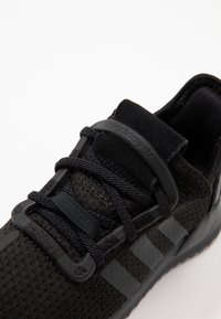 Svart atletisk sko med ovandel i mesh, platta skosnören och vadderad krage. Har tre strukturerade svarta ränder på sidan.