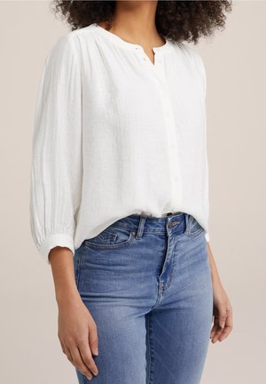 Femme portant un chemisier blanc à manches longues boutonné et un jean bleu taille haute sur un fond beige.