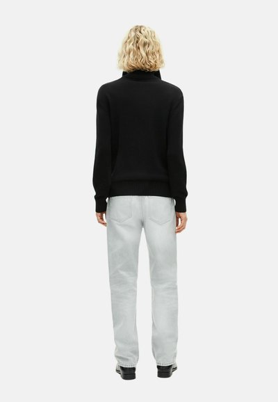 Personne blonde portant un pull noir à col roulé, un jean gris clair et des chaussures noires, se tenant de dos sur un fond blanc.