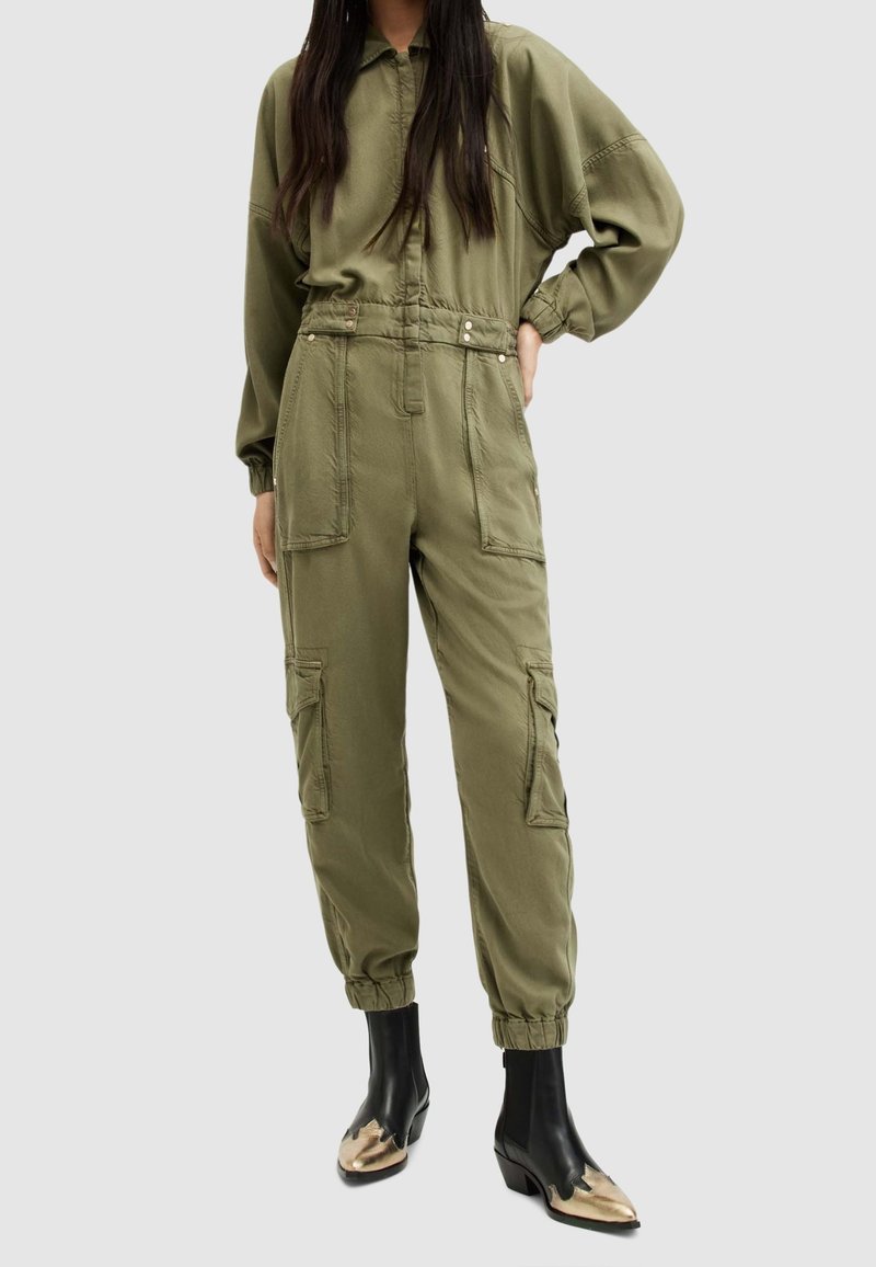 Femme portant une combinaison utilitaire vert olive avec de grandes poches et des poignets élastiques, associée à des bottines noires aux embouts métalliques dorés.