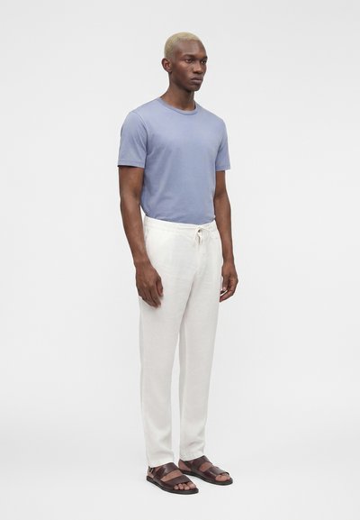 Homme aux cheveux blonds courts se tenant debout portant une chemise bleu clair à manches courtes, un pantalon blanc à cordon et des sandales marron foncé sur un fond uni.