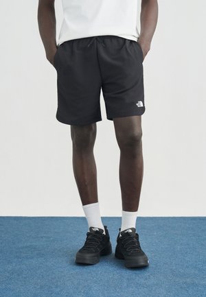 Homme portant un short noir The North Face, des chaussettes blanches, des baskets noires, debout les mains dans les poches sur un sol recouvert d'un tapis bleu.